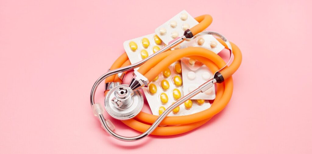 A flat lay photograph showing an orange stethoscope wrapped around yellow and white pills on a light pink background.