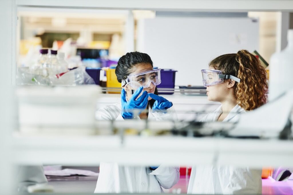 Two healthcare technicians stand behind a shelf in a clinical laboratory.