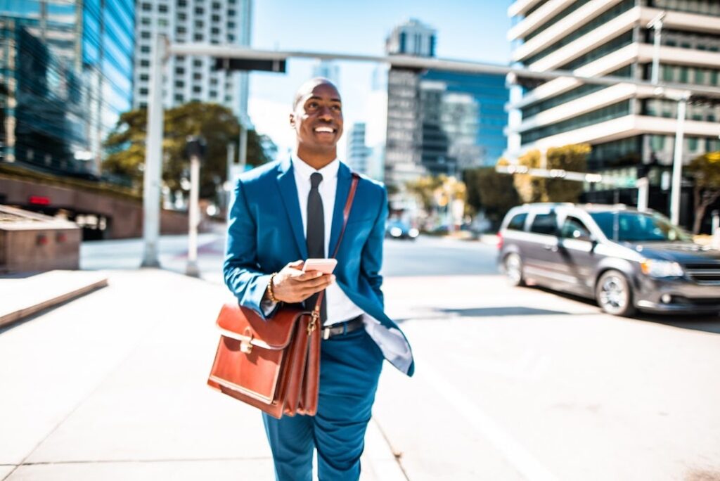 A man in a blue suit walks down the street, smiling while carrying a briefcase and cell phone.