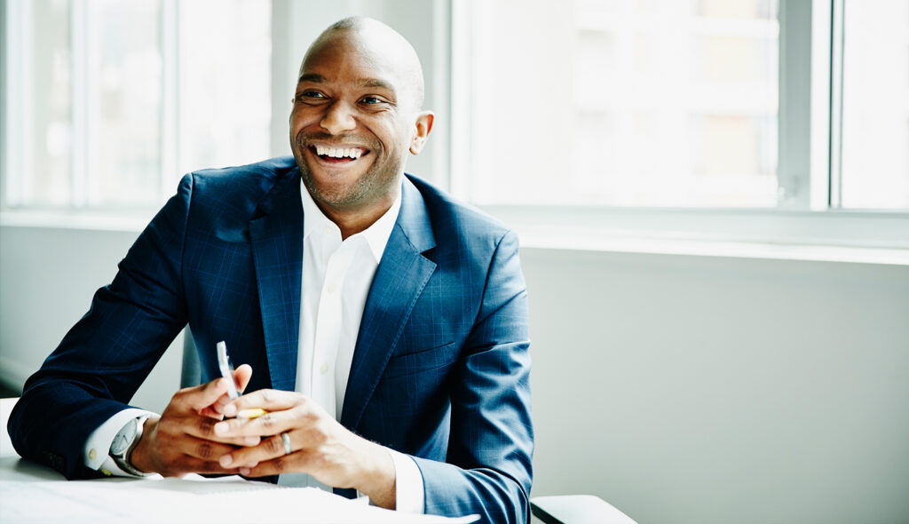 Seated in a conference room, a Black male healthcare MBA executive dressed in a white dress shirt and suit jacket holds a smartphone and smiles.