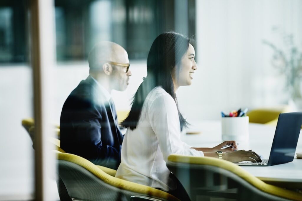 Two coworkers sit at a table in a conference room.