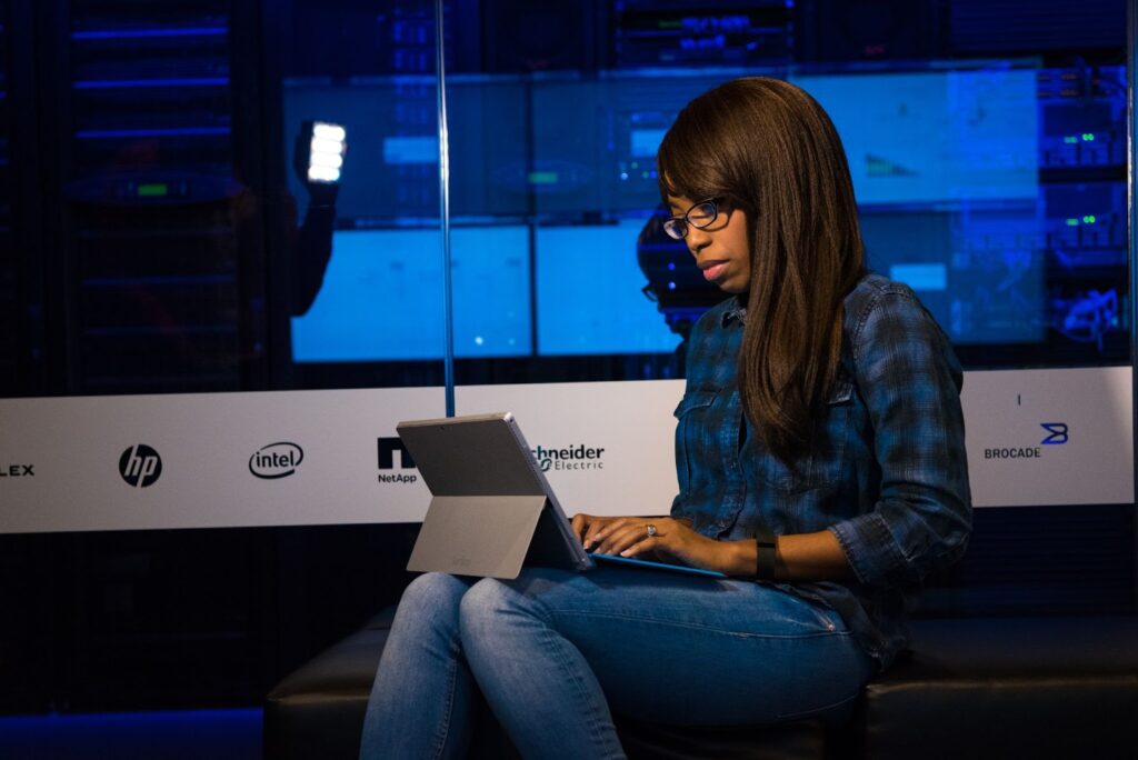 A Black woman wearing glasses sits and works at a computer laptop in a tech office.