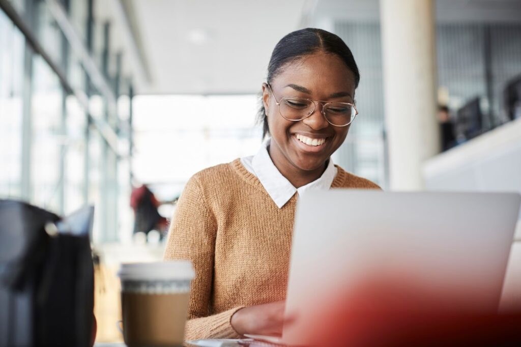 Seated at a workstation, a Black female computer science student in a brown sweater and white collared shirt works at a computer.