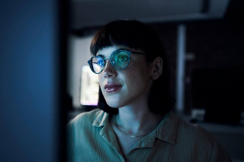 A woman works at a computer monitor, the screen content reflected in her eyeglasses.