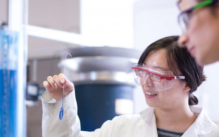 Two students wearing safety glasses and lab coats examine a component of a project.