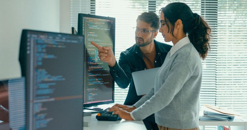 Two computing professionals stand before several large monitors displaying coding text; one points to a screen while the other types at a keyboard.